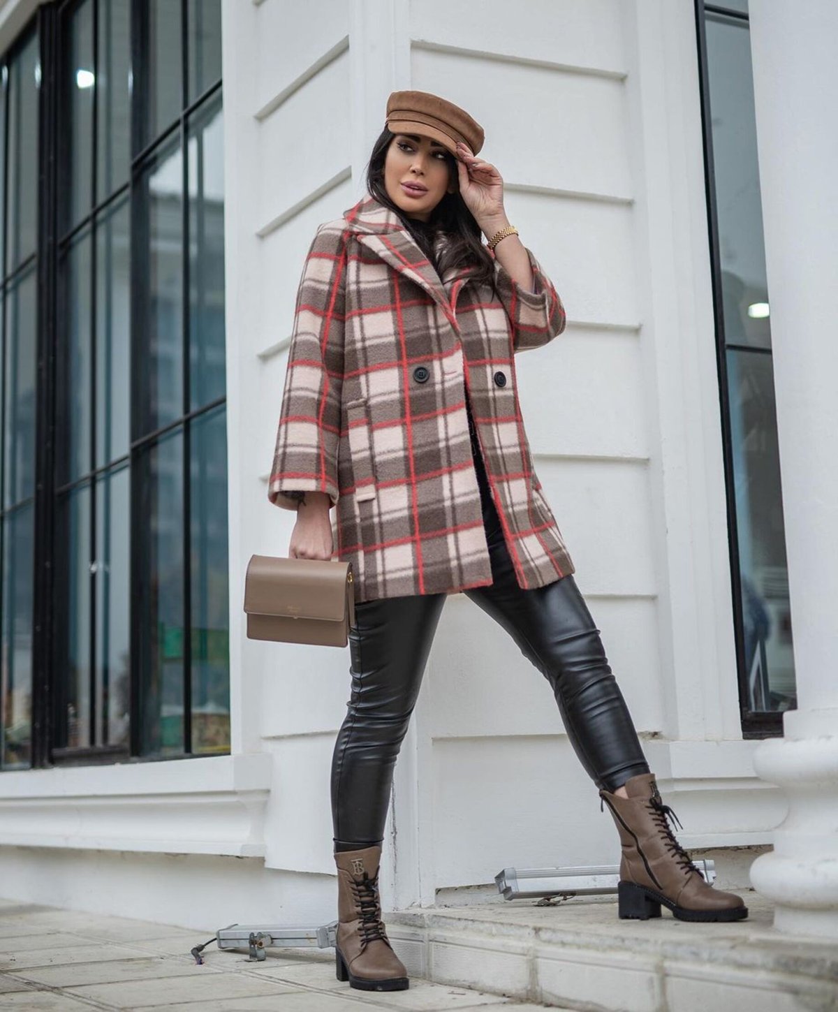 Woman in a beige plaid coat, flat cap and tan knee-high boots posing outdoors