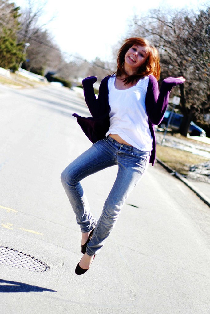 A woman mid-jump on a sunny street wearing an open long purple cardigan over a white t-shirt with faded blue skinny jeans and black ballet flats
