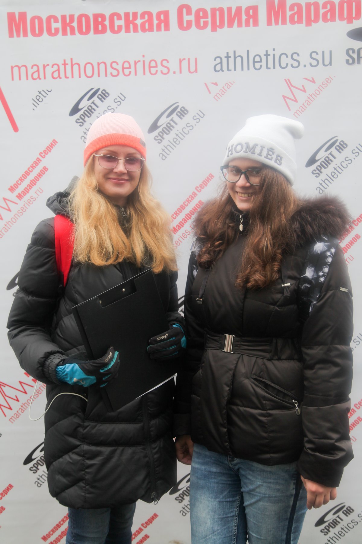 Two young women posing together wearing black quilted puffer jackets, blue jeans and knit beanies in coral and white