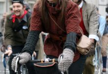 Woman riding an orange bicycle in a red and black checked tweed jacket, grey knit gloves, dark cap and a brown crossbody bag.