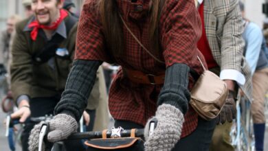 Woman riding an orange bicycle in a red and black checked tweed jacket, grey knit gloves, dark cap and a brown crossbody bag.