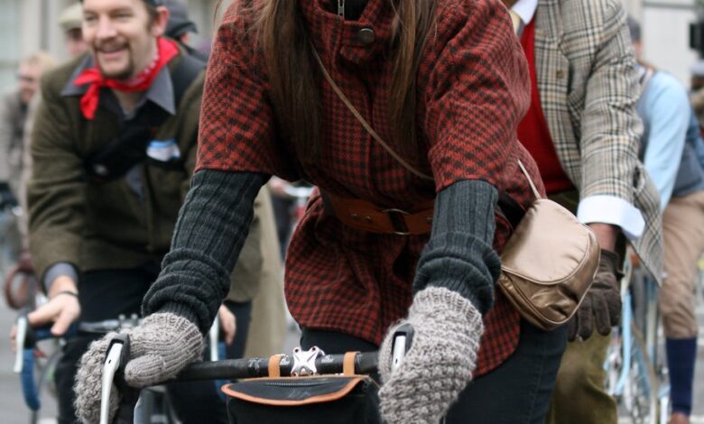 Woman riding an orange bicycle in a red and black checked tweed jacket, grey knit gloves, dark cap and a brown crossbody bag.