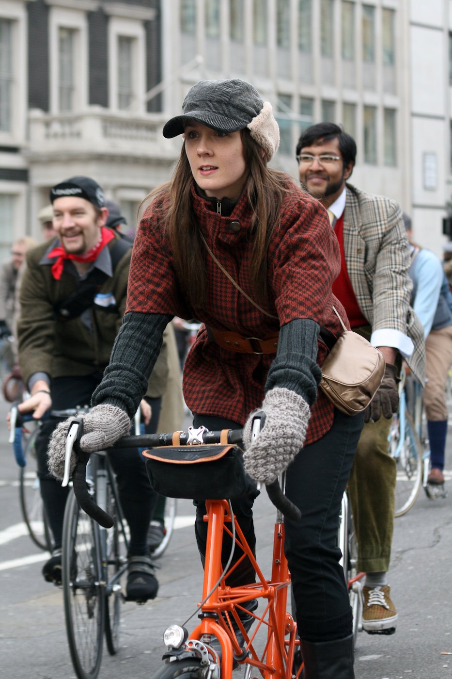 Woman riding an orange bicycle in a red and black checked tweed jacket, grey knit gloves, dark cap and a brown crossbody bag.