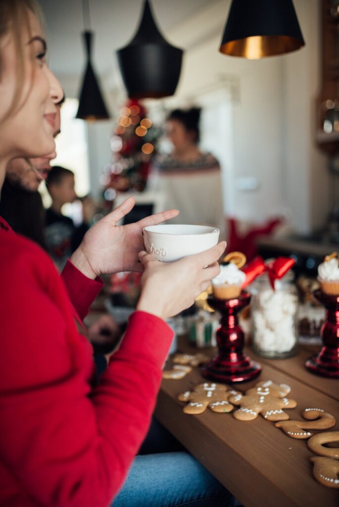 A smiling woman in a red Christmas sweater holding a decorated holiday mug at a Christmas table with friends