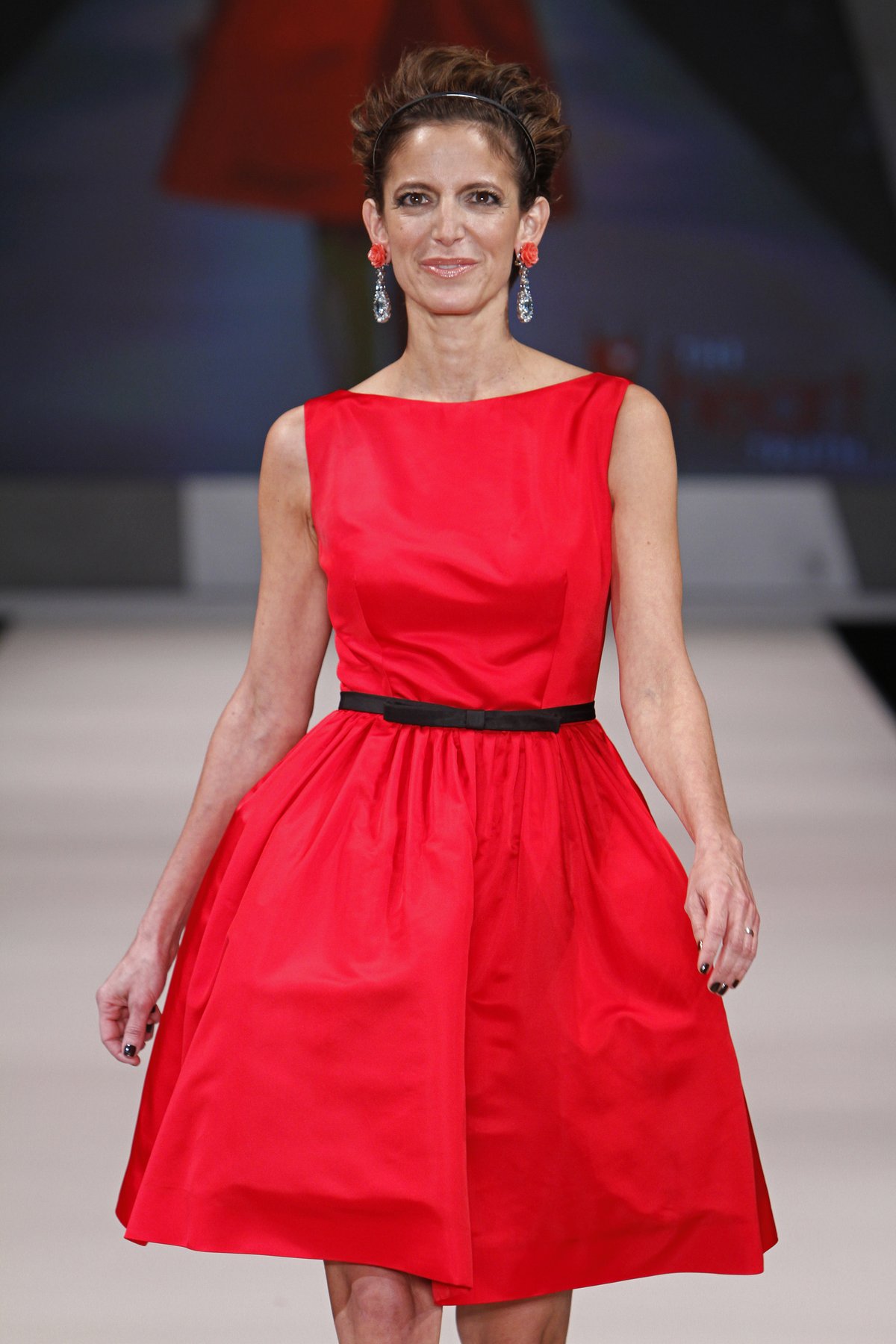 A woman walking a runway in a red fit-and-flare midi dress with a thin black waist belt and a full skirt.