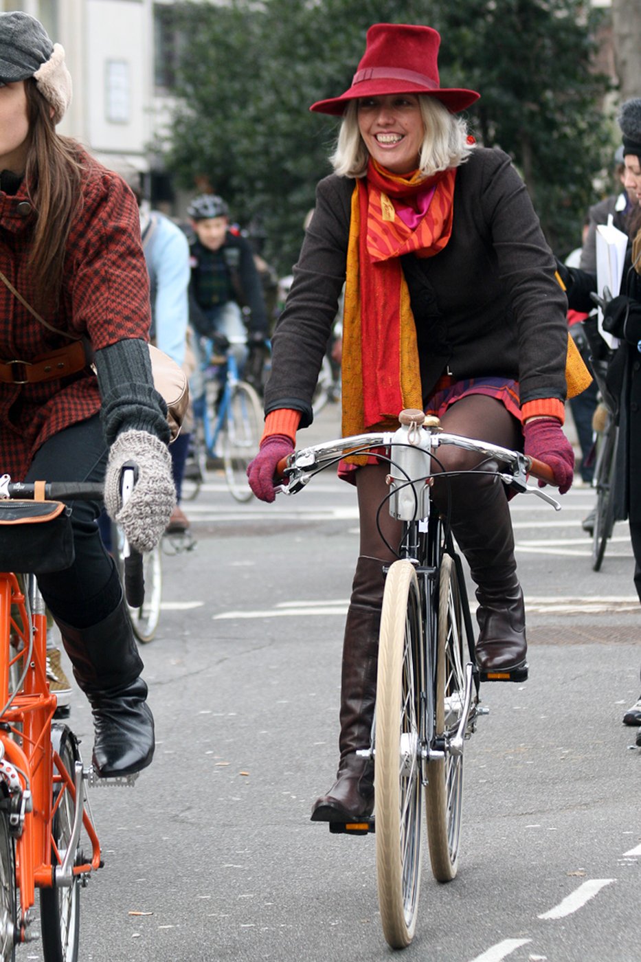 Smiling blonde woman in a red hat and orange-red scarf riding a bicycle, wearing a red plaid mini skirt, dark tights and knee-high boots.