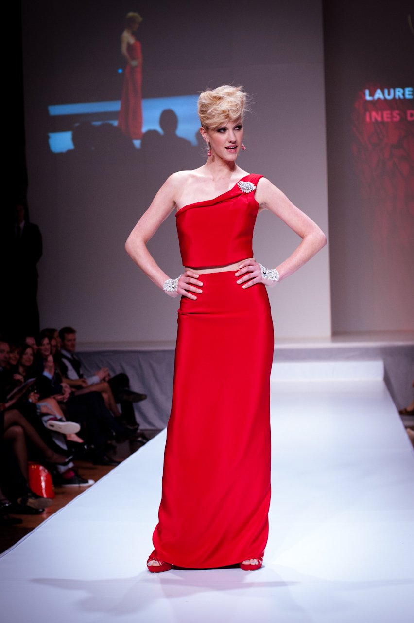 Model on a runway wearing a red one-shoulder two-piece evening gown with a crystal brooch, statement cuffs and red heels