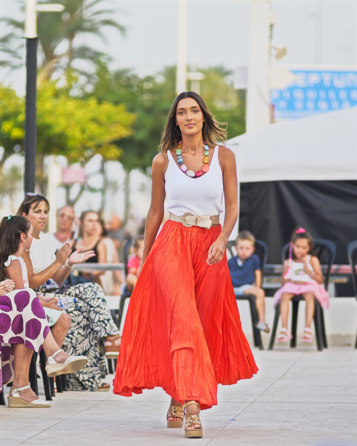 Model walking an outdoor runway wearing a white tank top with flowing red-orange pleated palazzo trousers, a wide woven belt and strappy platform sandals.
