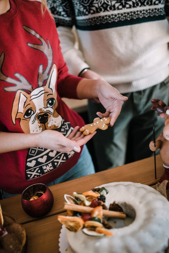 A woman in a red Christmas sweater with a cartoon reindeer riding a bulldog, holding a gingerbread man cookie at a holiday party table