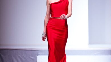 Woman walking a runway in a red satin one-shoulder crop top and matching long red satin skirt with a crystal brooch at the shoulder