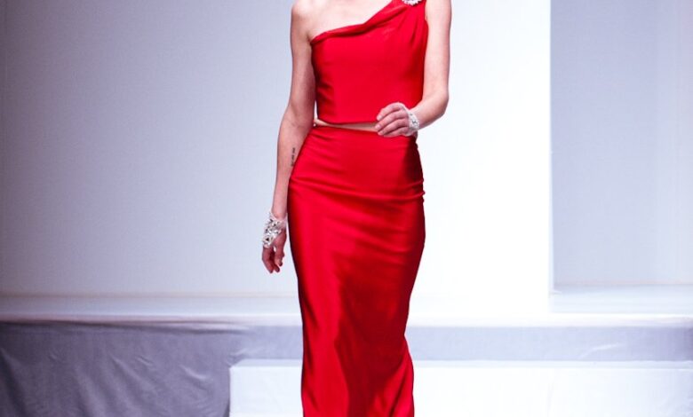 Woman walking a runway in a red satin one-shoulder crop top and matching long red satin skirt with a crystal brooch at the shoulder
