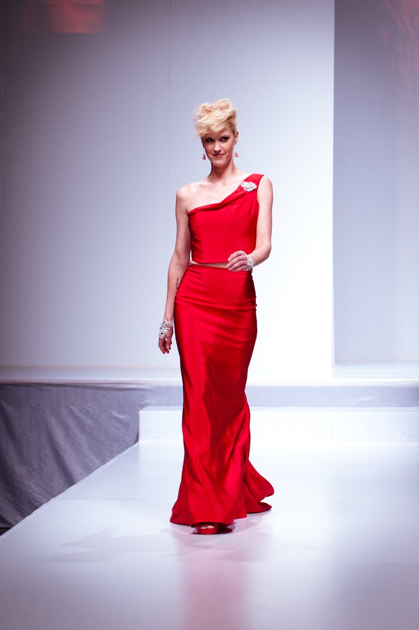 Woman walking a runway in a red satin one-shoulder crop top and matching long red satin skirt with a crystal brooch at the shoulder