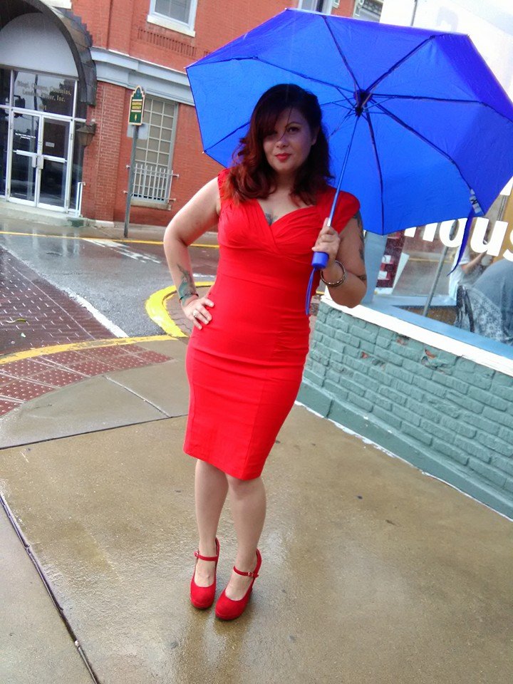 Woman in a red sheath dress and red Mary Jane heels holding a blue umbrella on a rainy street.