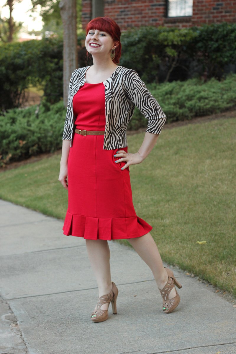 A woman with red hair wearing a red sheath dress with a ruffled hem, a zebra-print cardigan, a tan belt and tan cutout heels, standing on a sidewalk