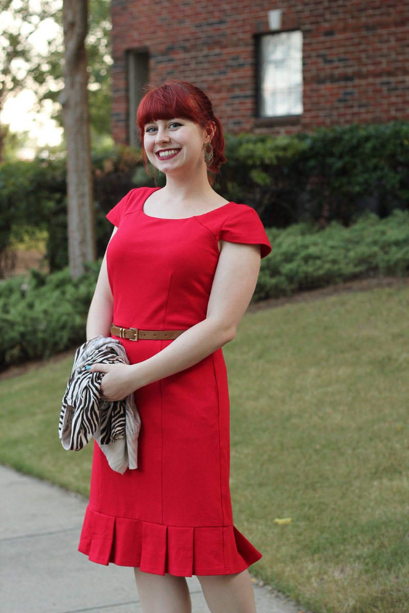 A smiling red-haired woman in a bright red sheath dress with a tan belt, holding a zebra-print scarf outdoors by a brick building