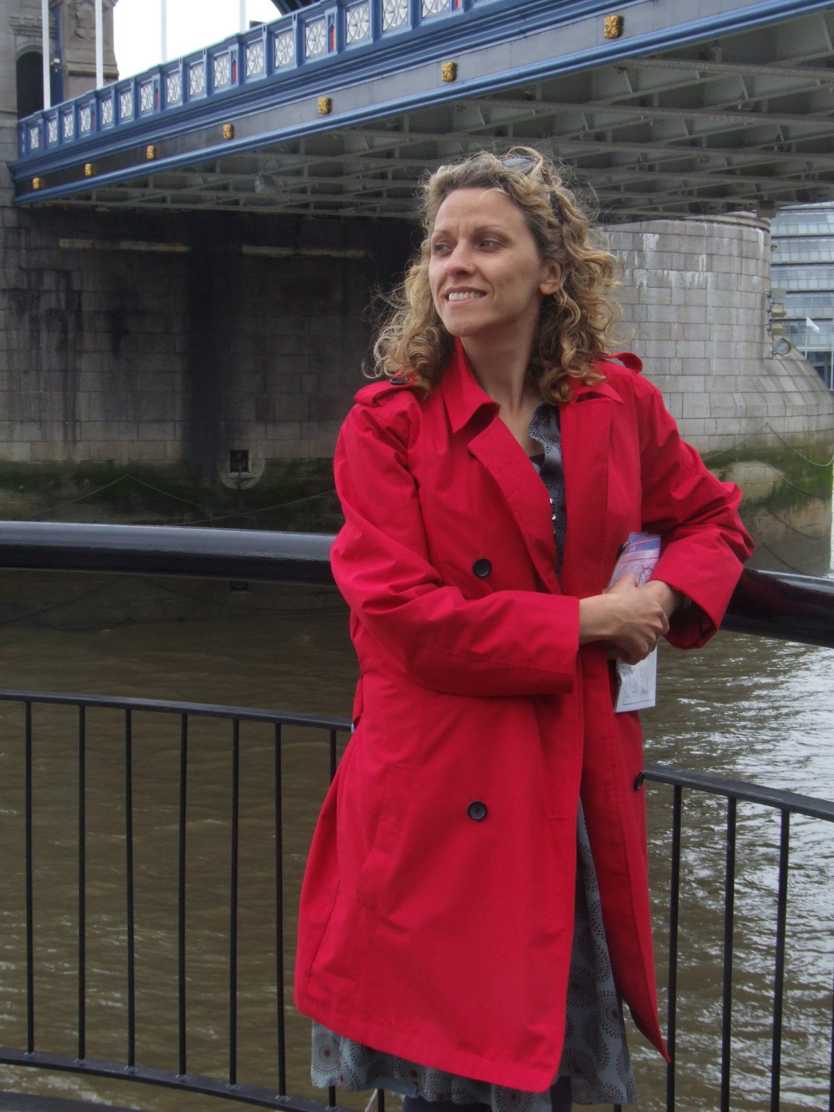 A smiling woman in a red double-breasted trench coat with a patterned dress underneath, standing by a railing with Tower Bridge behind her
