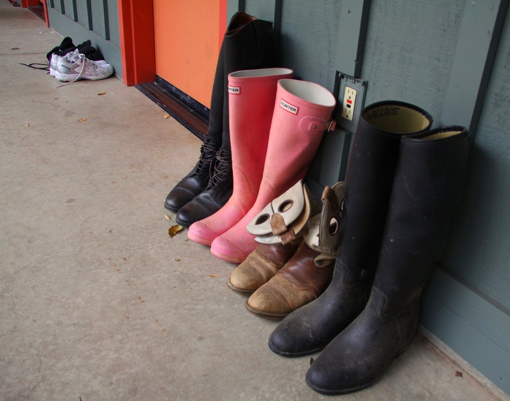 Color photograph of a row of tall boots by a stable door: black riding boots, pink wellington boots and brown lace-up boots