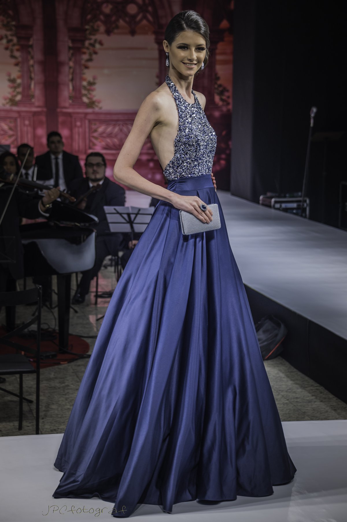 Model on a runway wearing a royal blue halter evening gown with a sequin bodice and full satin skirt, holding a silver clutch