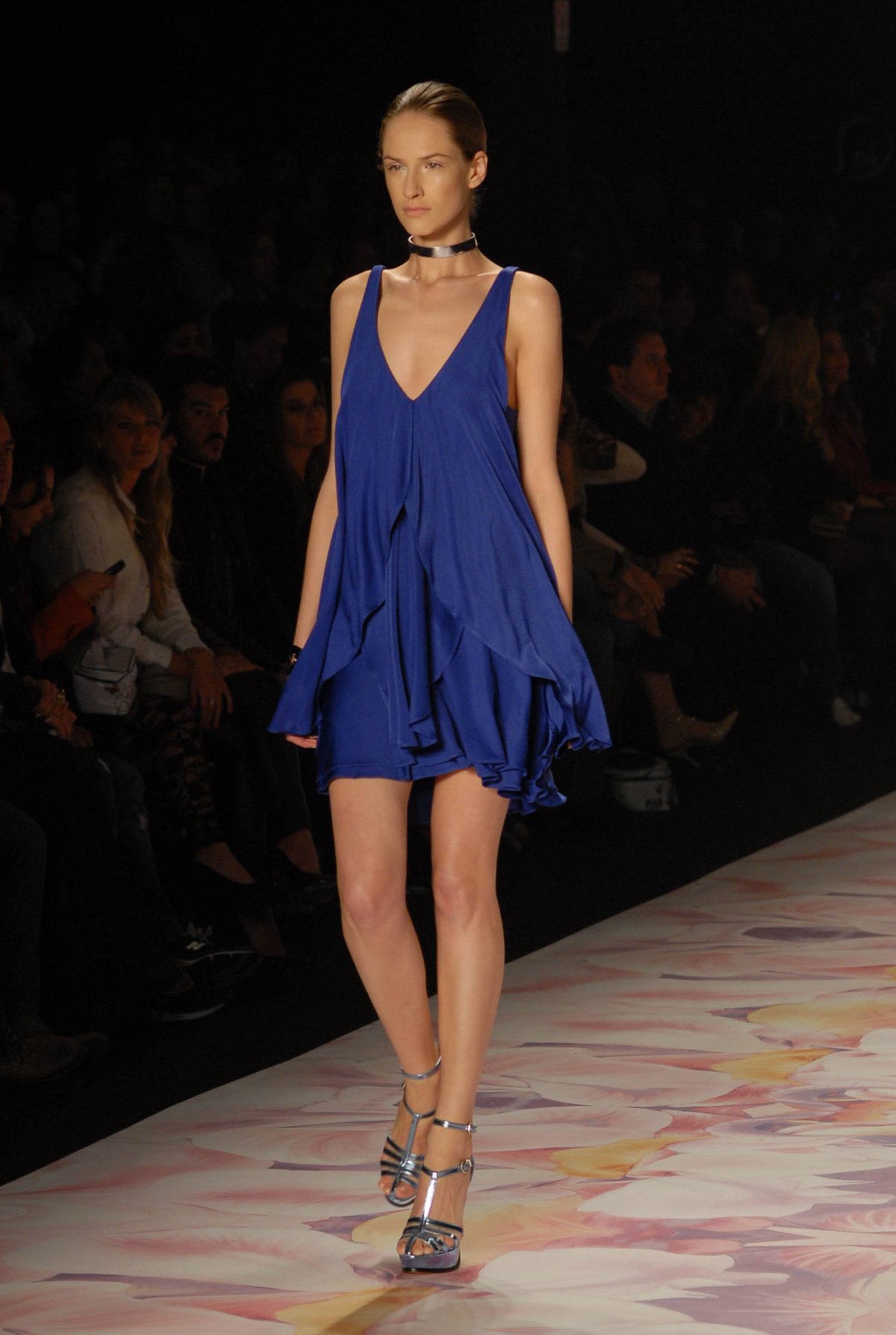 Model on a runway wearing a short royal-blue ruffled cocktail dress with a choker necklace and strappy heels