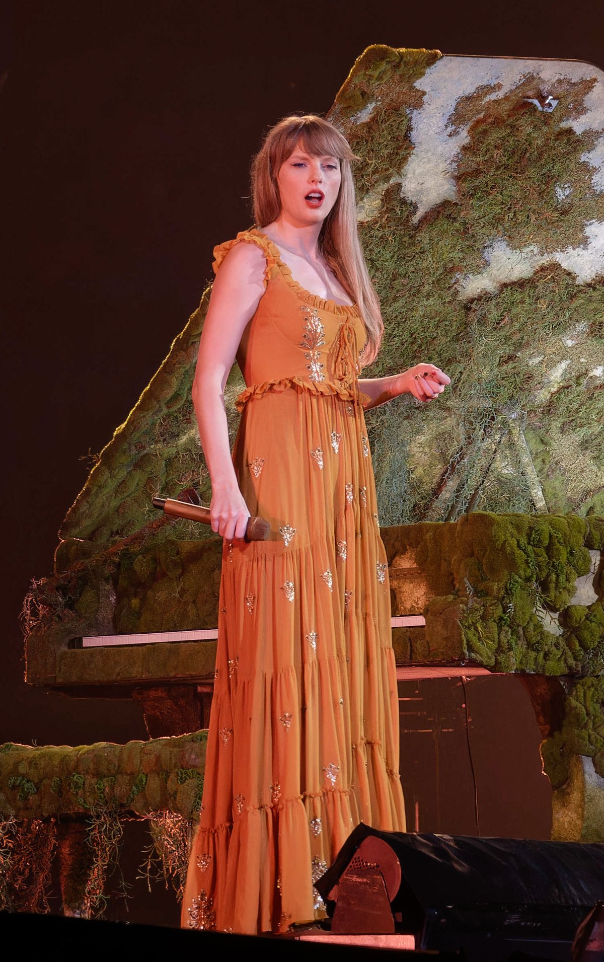 Blonde performer on stage wearing a rust-caramel tiered maxi dress with gold embroidery and a lace-up neckline, beside a moss-covered piano.