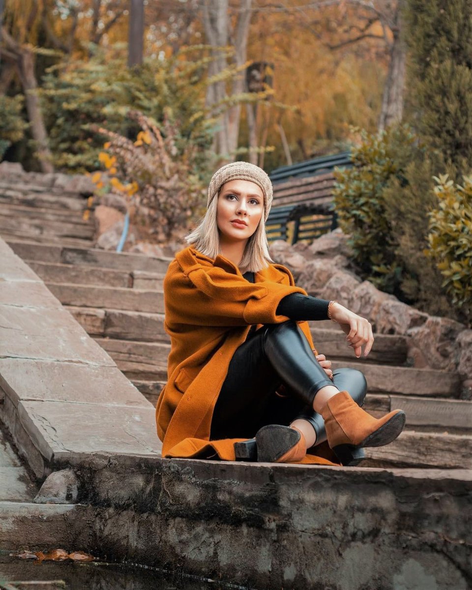 A blonde woman in a rust-orange wool coat, black knit top, leather leggings and tan ankle boots with a knit beanie, seated on stone steps in an autumn park