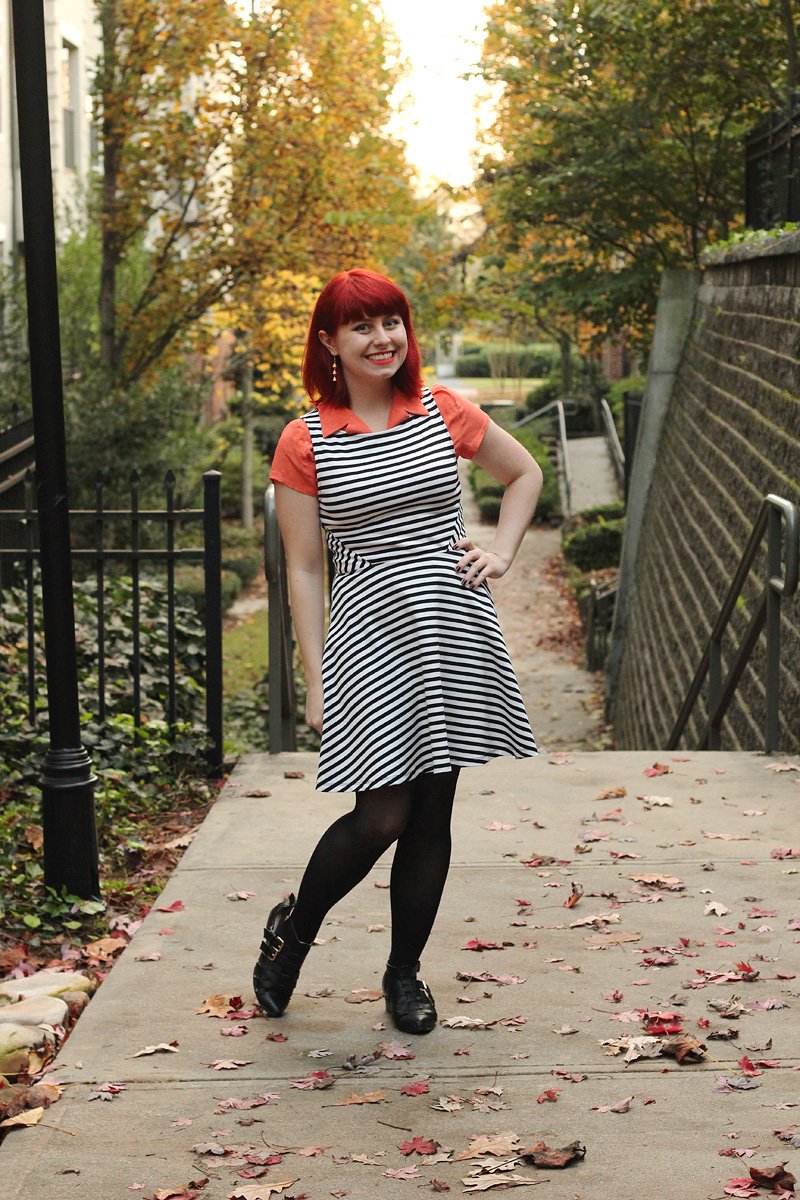 Red-haired woman on an autumn path strewn with leaves wearing a black-and-white striped dress over a coral-orange collared shirt with black tights and black buckled ankle boots.