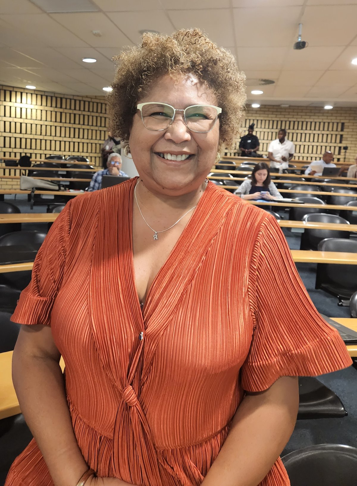 Smiling curly-haired woman with glasses wearing a rust-terracotta pleated v-neck top with flutter sleeves and a front knot.