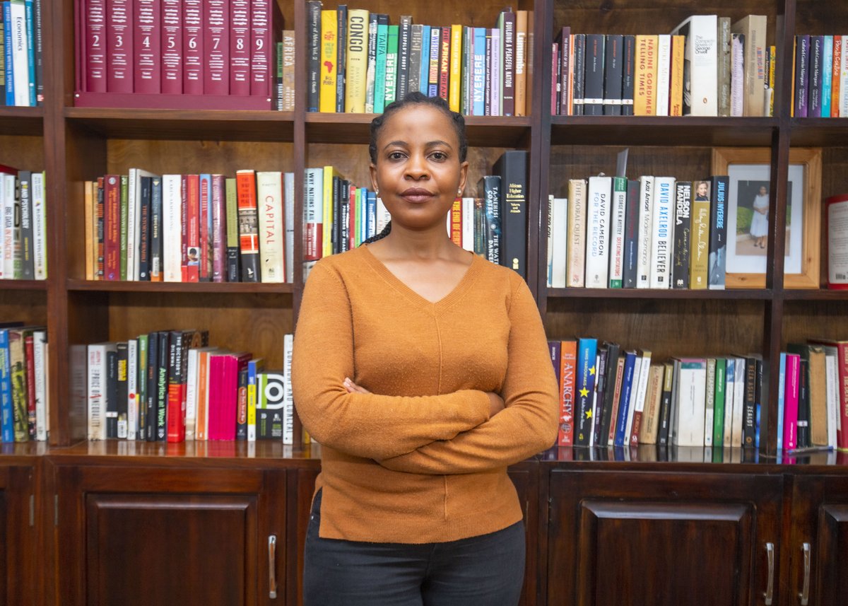 Woman with arms crossed in front of bookshelves wearing a rust-copper v-neck knit sweater with dark trousers.