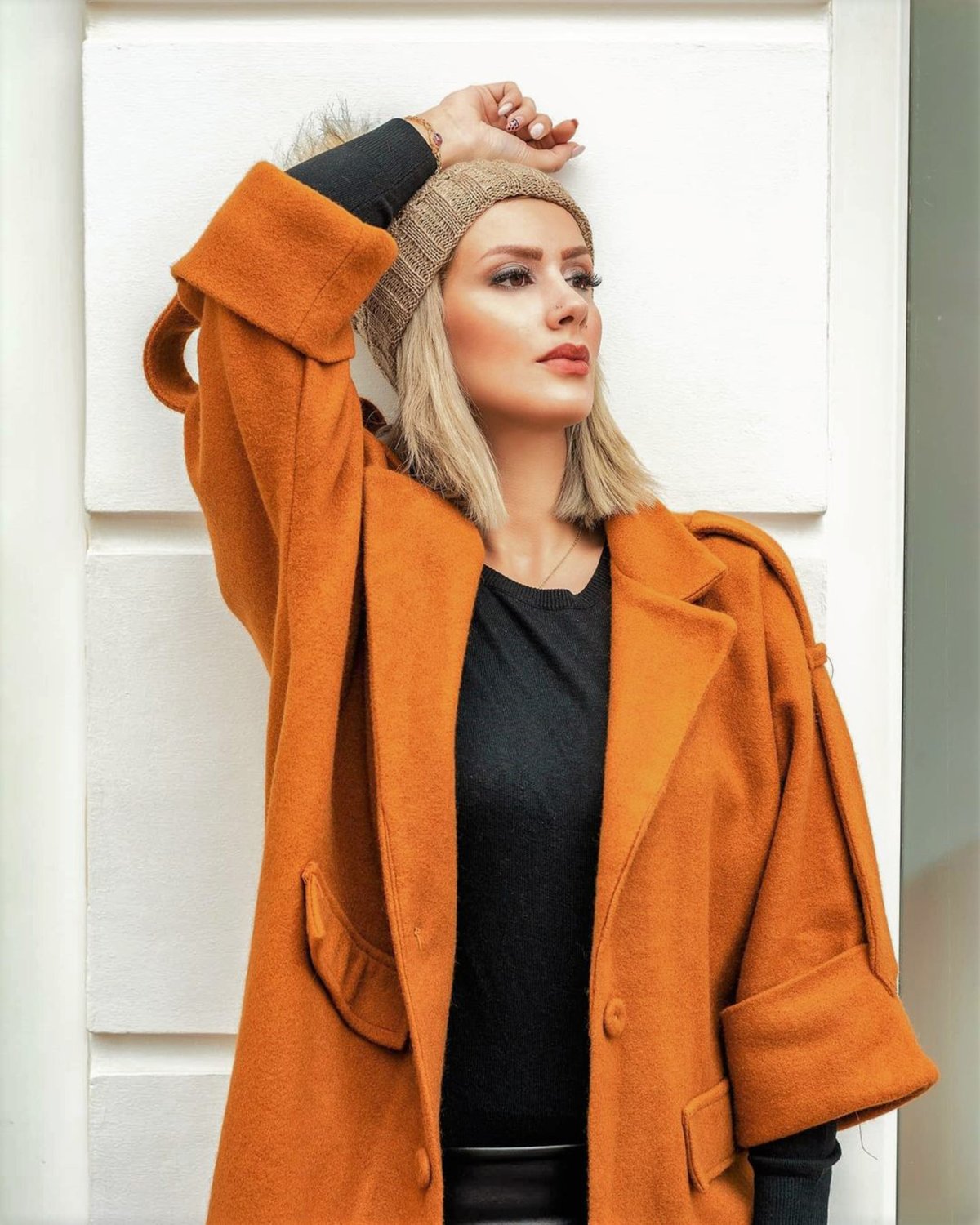 Woman in a rust wool coat and beige knit beanie leaning against a white wall