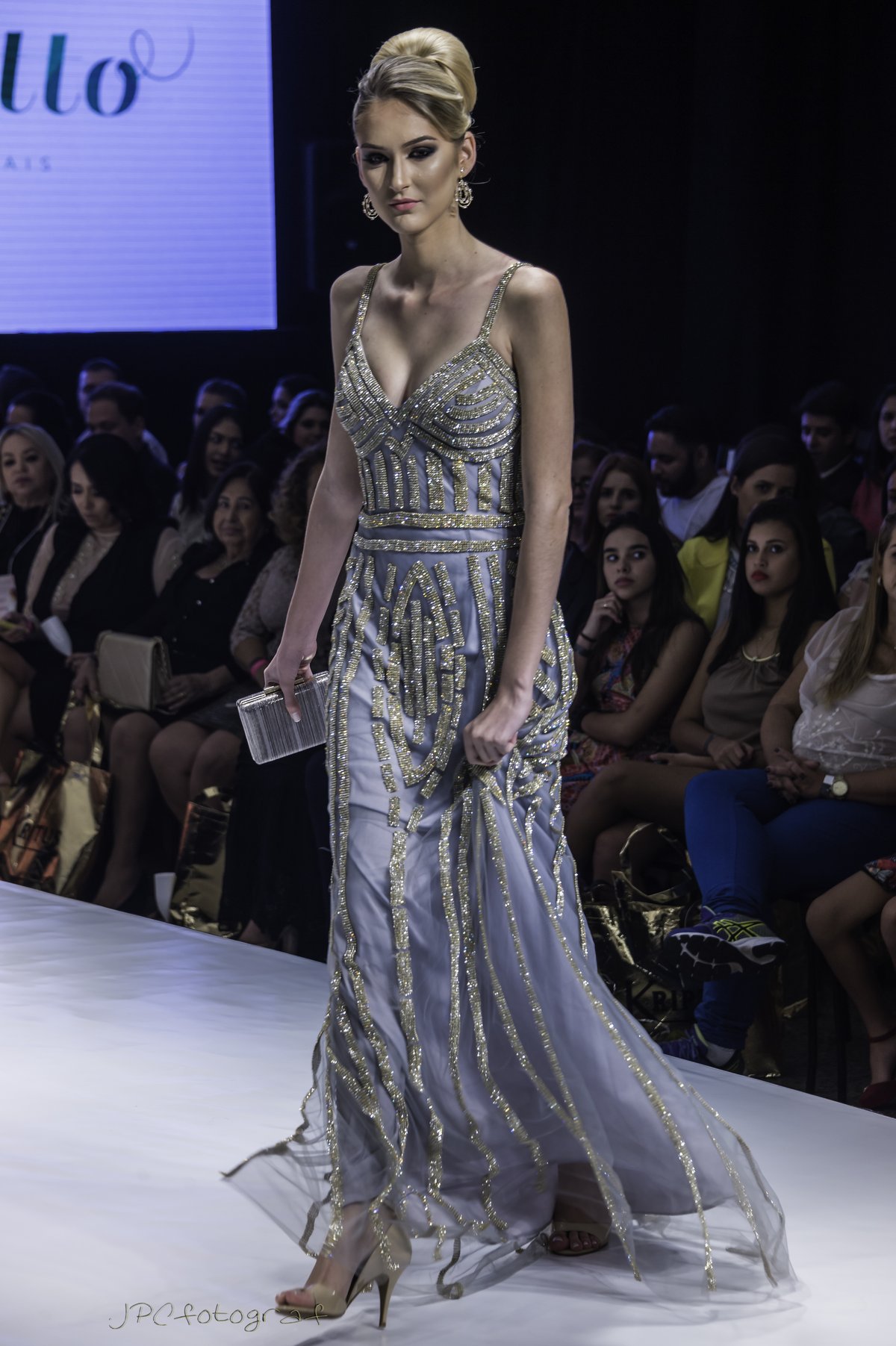 Model on a runway wearing a silver-grey evening gown with vertical gold bead stripes, thin straps and a V-neckline