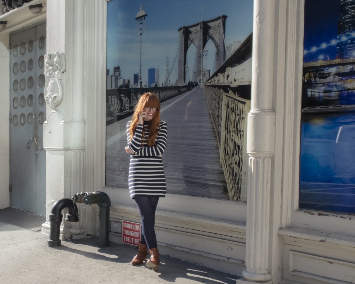 A red-haired woman on a New York sidewalk wearing a navy-and-white striped knit mini sweater dress with navy tights and tan ankle boots, talking on a phone in front of a Brooklyn Bridge mural.