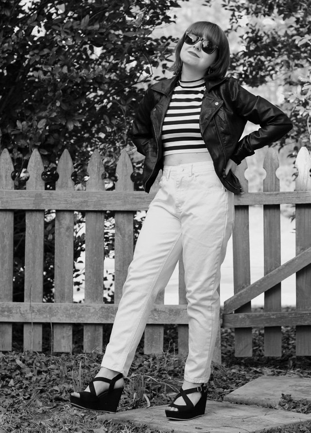 Black-and-white full-length photo of a woman with a bob haircut and sunglasses standing beside a wooden picket fence, wearing a black-and-white horizontally striped cropped mock-turtleneck top, an open black leather biker jacket, white high-rise jeans, and black platform wedge sandals.