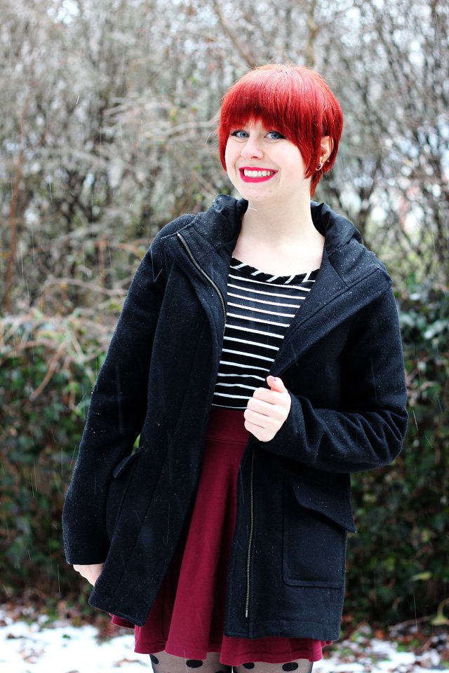 A woman with a red pixie cut wearing a black-and-white striped top under a black wool coat with a dark maroon skirt and polka-dot tights, standing in a park