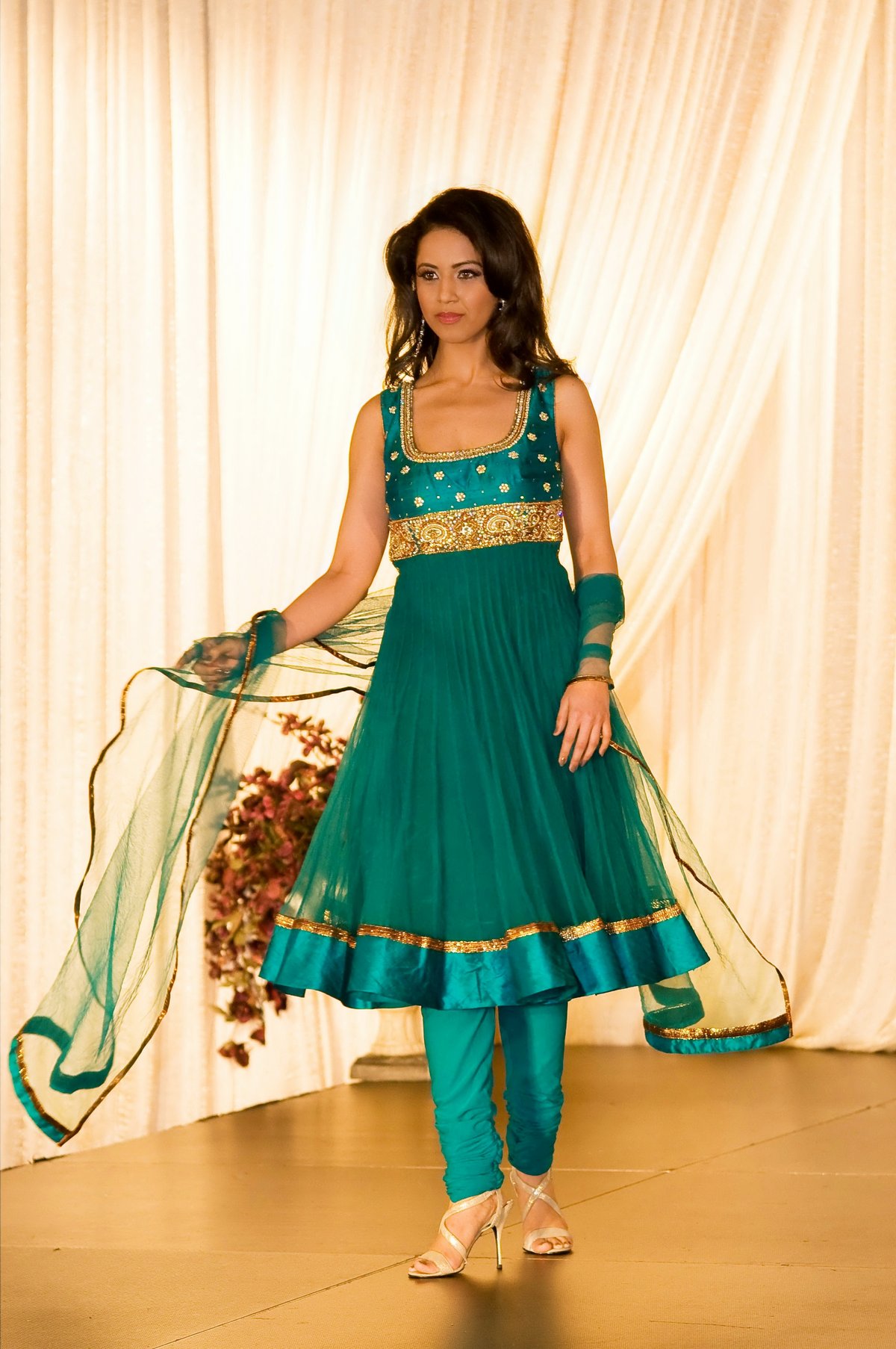 Model walking a fashion show runway in a sleeveless teal green anarkali with a gold embroidered bodice, matching churidar and sheer dupatta