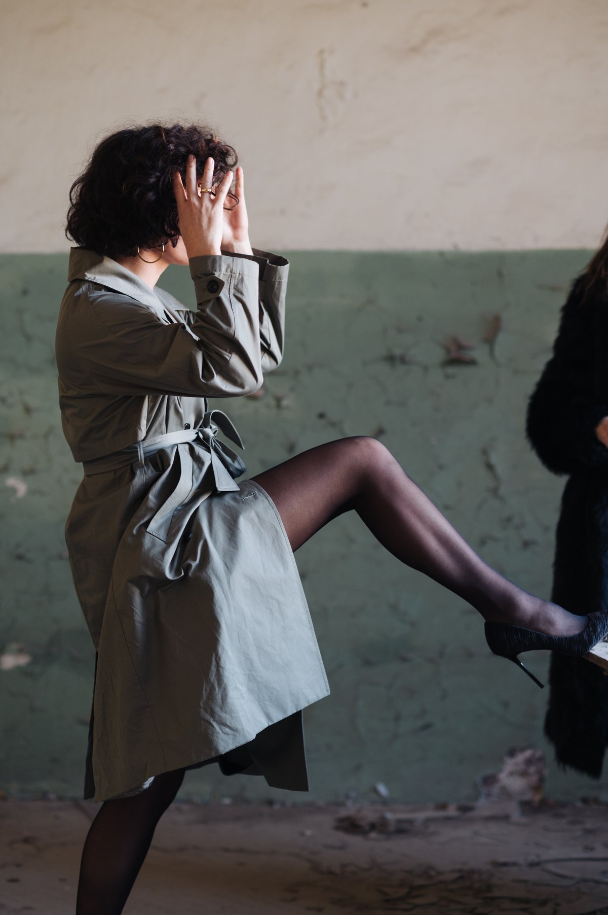 Editorial fashion photograph of a woman in a belted olive-toned trench coat with sheer black tights and black stiletto heels, one leg raised on a ledge