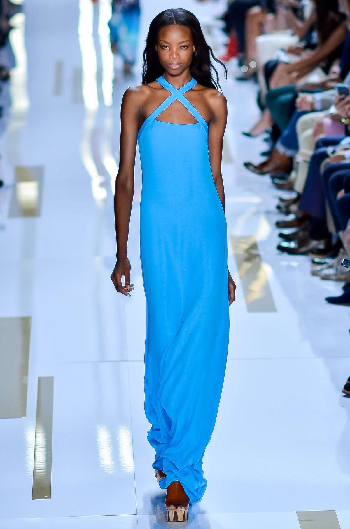 Model on a runway wearing a bright turquoise-blue maxi dress with a crisscross halter neckline