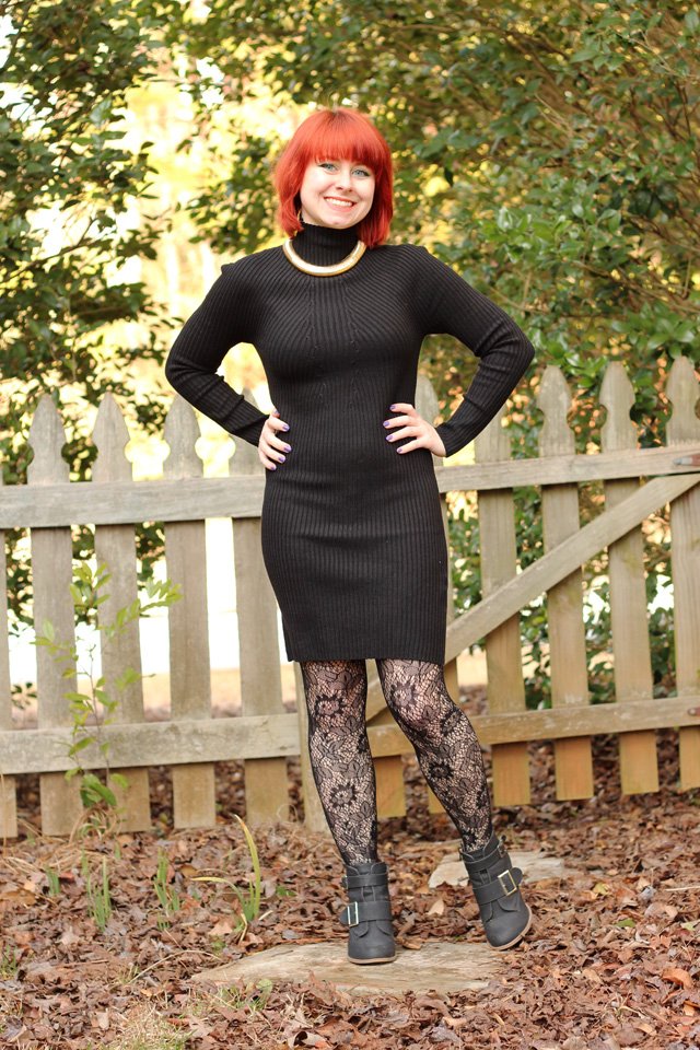 A woman with red hair wearing a black turtleneck sweater dress, black lace tights and grey ankle boots, standing by a wooden fence
