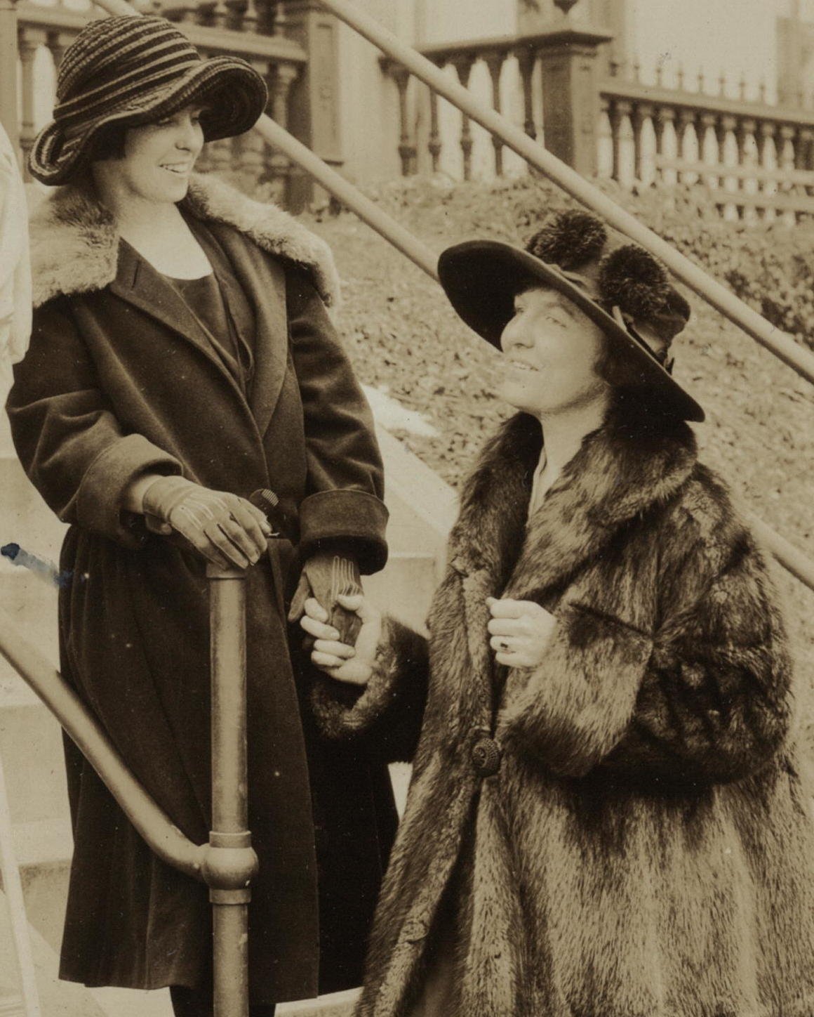 A sepia photo of two women chatting on outdoor steps in the 1920s, one in a long dark fur coat with a fur collar and the other in a dark wool coat with a light fur collar, both wearing wide-brimmed hats and gloves.