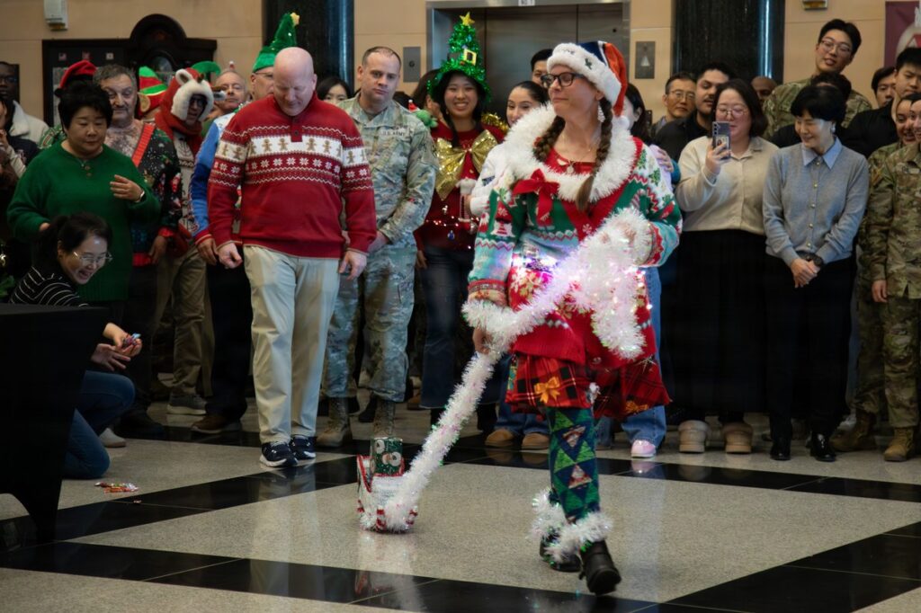 A woman walking at an ugly sweater contest wearing a sweater draped in tinsel, lights and a Santa hat with a tartan skirt