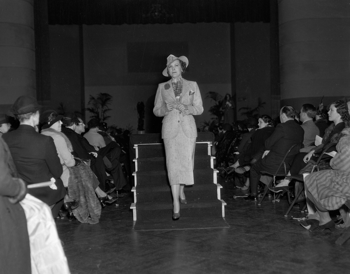 Black-and-white photo of a woman descending runway stairs in a light-toned tweed skirt suit, striped scarf and hat, at a 1936 Los Angeles fashion show.
