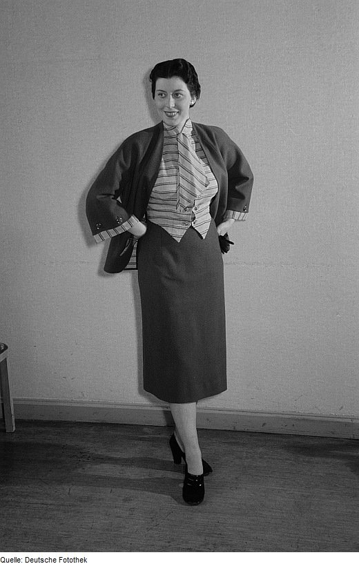 Vintage black-and-white photograph of a woman in a dark skirt suit with a striped blouse, hands in pockets