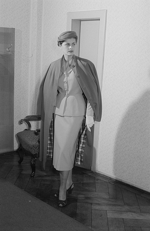 Vintage black-and-white photograph of a woman in a light-coloured skirt suit with a coat draped over her shoulders, wearing a cap and gloves
