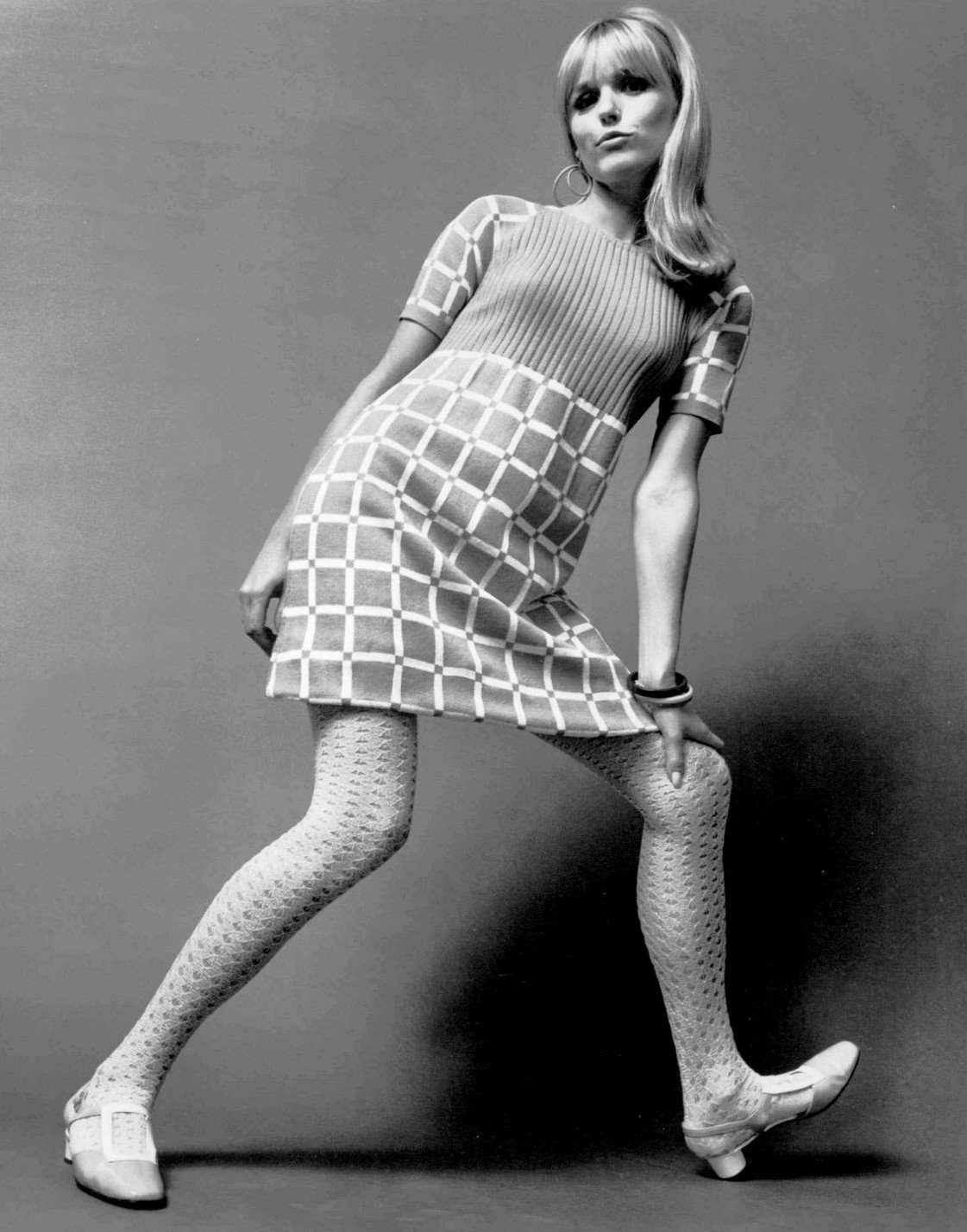 Black-and-white 1967 studio photograph of a model wearing a ribbed, geometrically patterned sweater-knit dress with patterned tights and chunky shoes.
