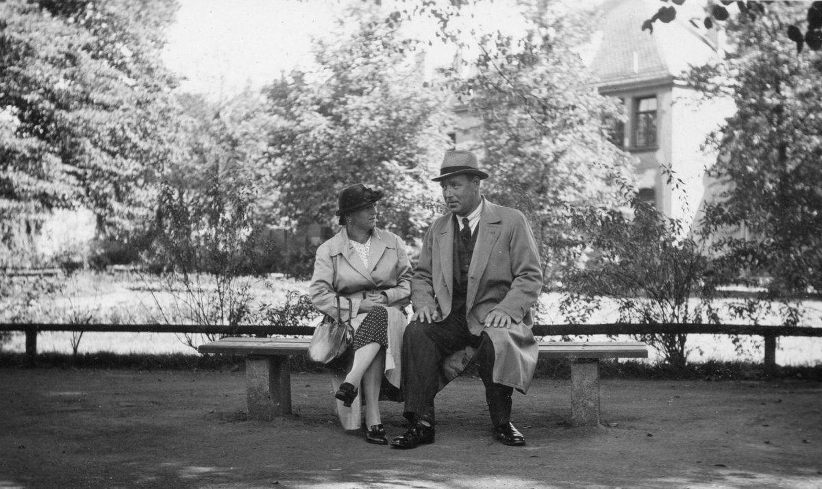 Black-and-white Fortepan photograph of a woman in a light trench coat and dark hat sitting on a park bench beside a man in a long coat