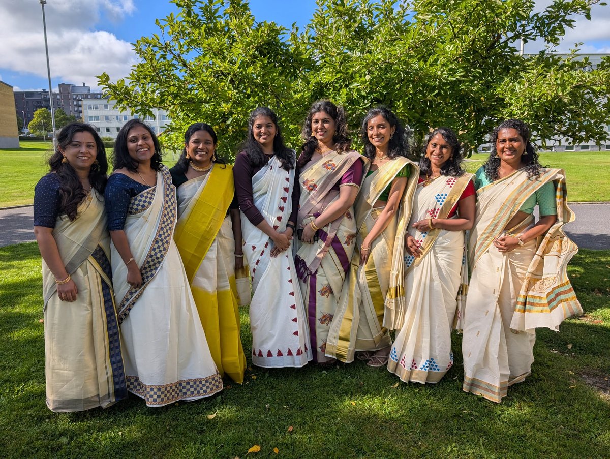 Eight smiling women standing in a park wearing white and off-white Kerala-style sarees with gold and coloured borders, trees and a building behind them