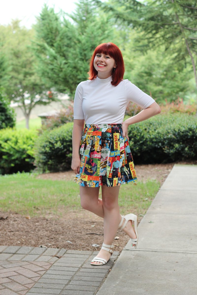 Smiling young woman with red hair standing on a park path wearing a white mock-neck top with a clearly visible high collar, a colorful comic-print pleated skater skirt, and white strappy flat sandals.