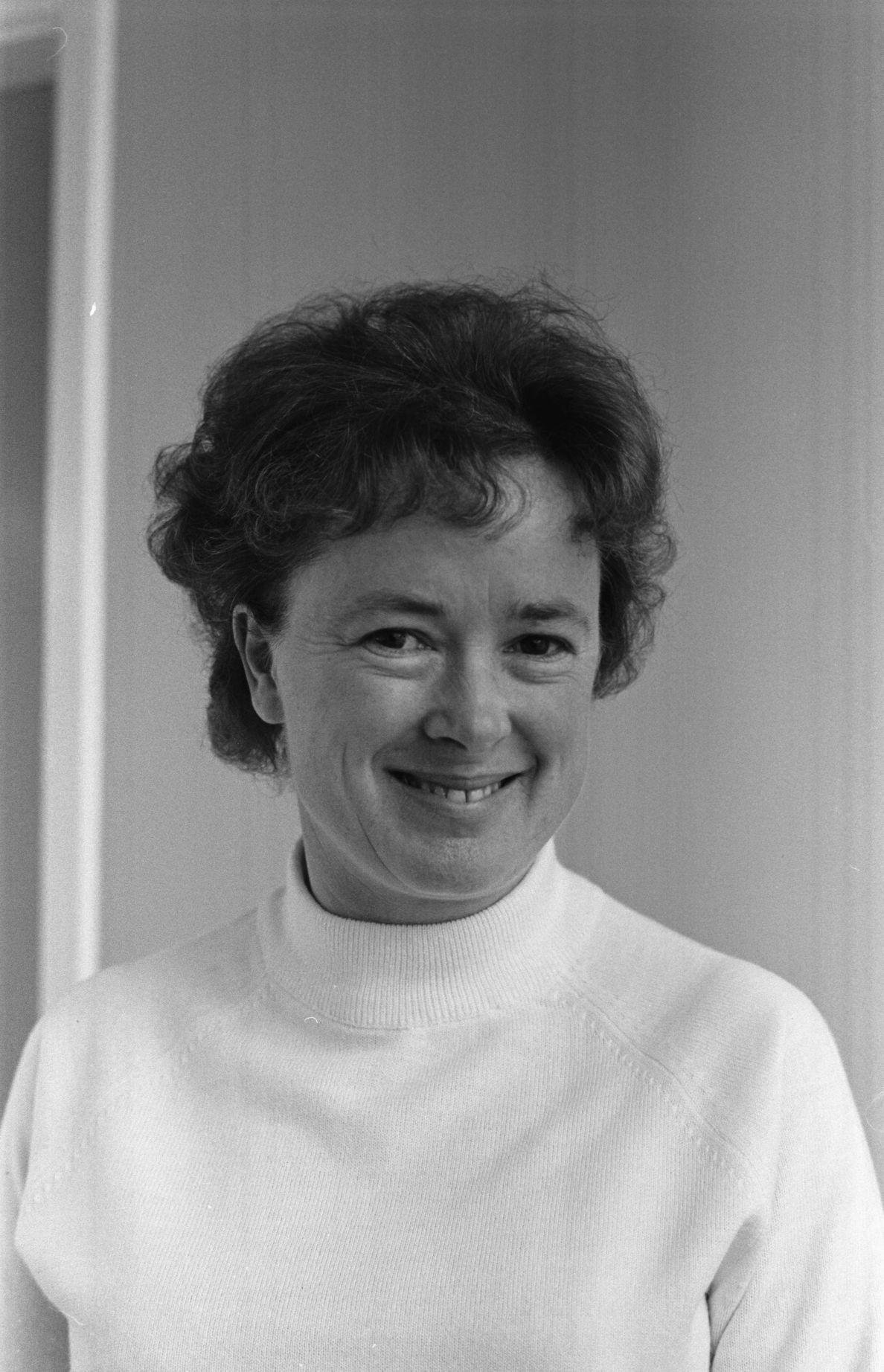 Black-and-white vintage portrait of a smiling woman with dark curly hair wearing a white ribbed polo-neck jumper, the high collar visible at her neck.
