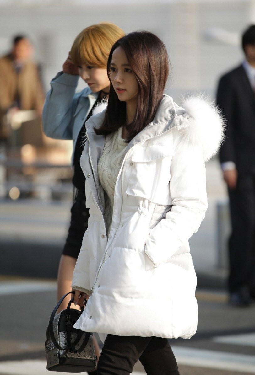 A young woman walking outdoors wearing a white quilted puffer parka with a white fur-trim hood over a cream cable-knit sweater, black skinny pants, and carrying a black handbag