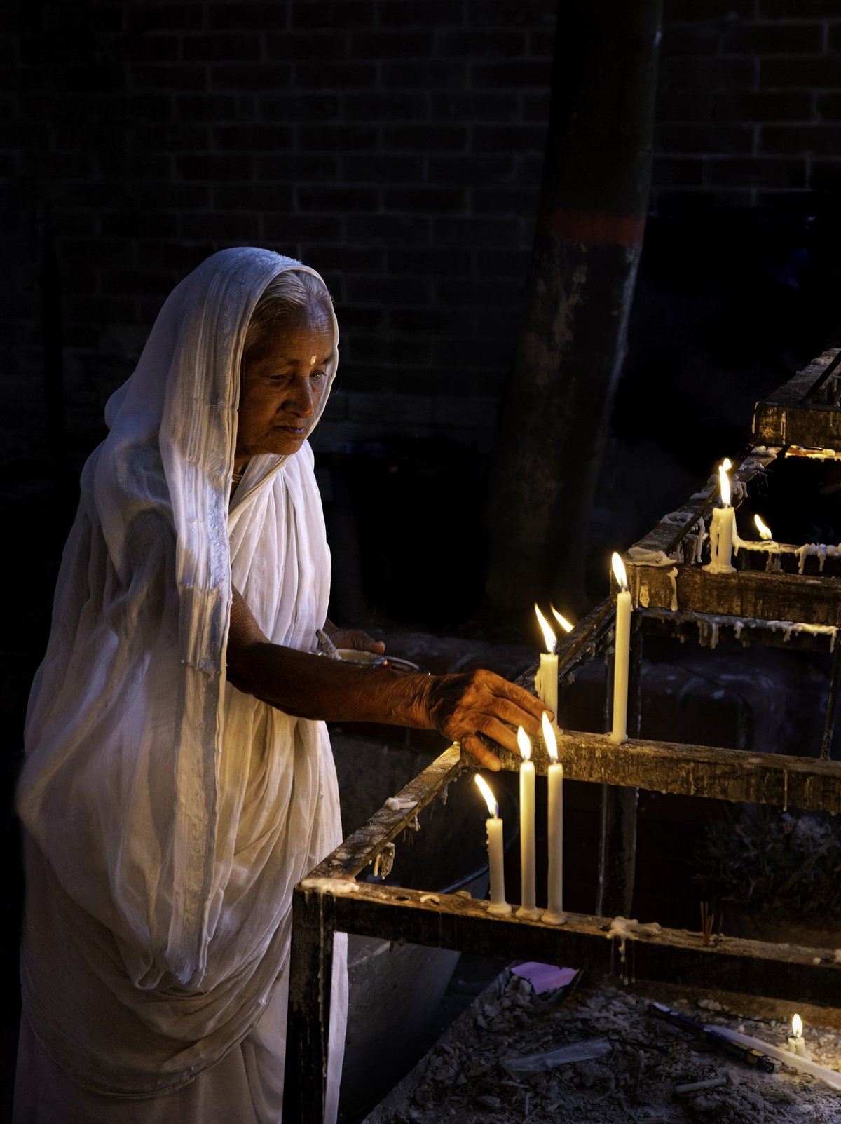 An elderly woman in a plain white saree with her head covered, lighting candles at a candle stand in a dark temple