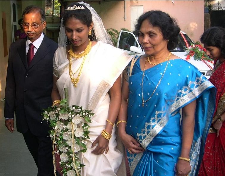 A bride in a white saree with a veil, gold necklaces and bangles, holding a white bouquet, walking with family at a wedding